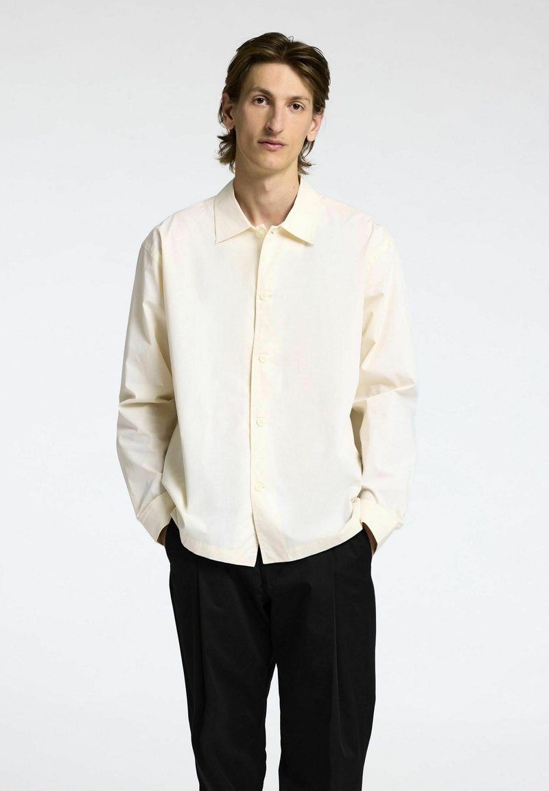 Camicia button-up color crema con colletto a punta, maniche lunghe e vestibilità ampia, abbinata a pantaloni neri. Tessuto morbido.