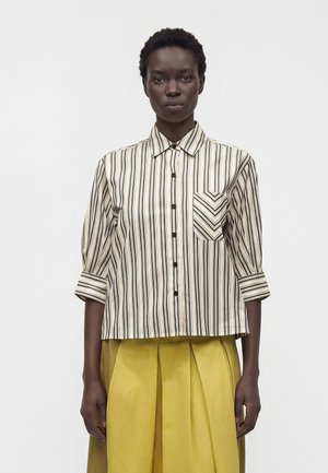 Femme portant une chemise beige et noire à rayures verticales avec manches retroussées et une jupe plissée jaune, debout sur un fond blanc.