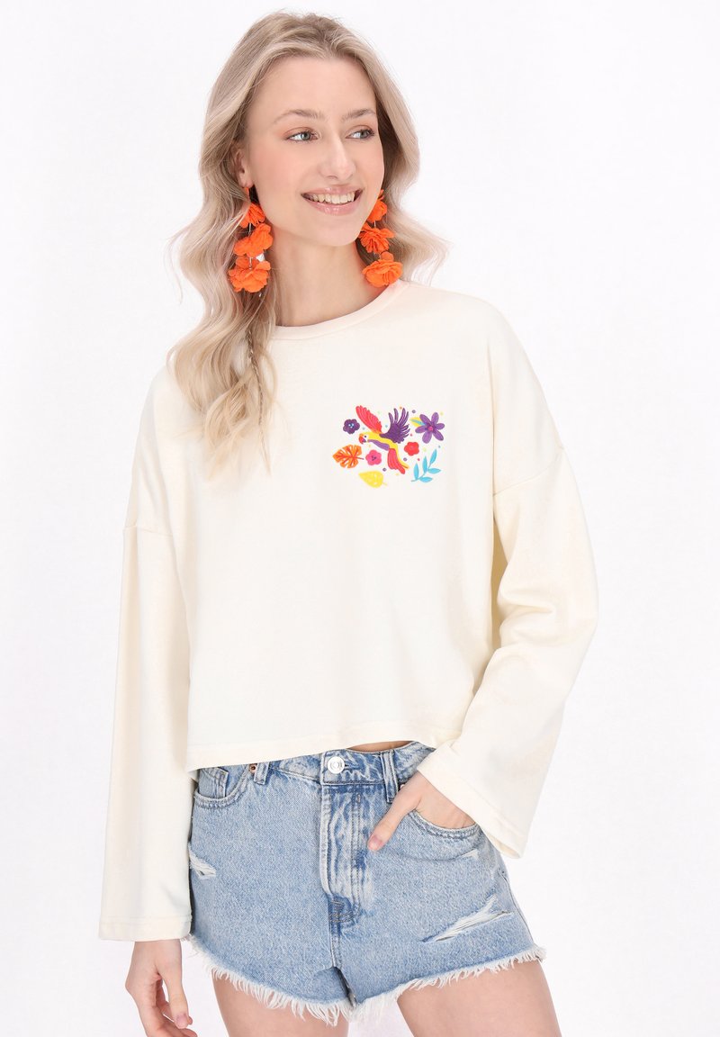 Sudadera de gran tamaño en color crema con bordado floral en colores vibrantes, combinada con shorts de mezclilla desgastados. El tejido parece suave y texturizado.