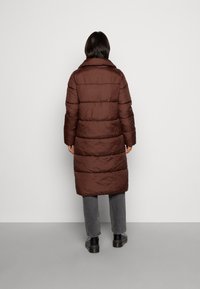 Langer brauner Puffermantel mit Stehkragen, gestepptem Design und einer leicht glänzenden Textur. Trägt er über grauen Hosen und schwarzen Stiefeln.