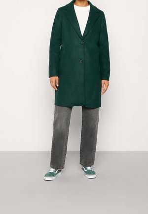 Person iført en mørkegrøn jakke med knapper over en hvid top, grå jeans med lige ben og grønne og hvide sneakers, stående på en grå gulv.