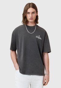 T-shirt gris oversize avec un col rond, des manches courtes et un logo blanc sur la poitrine gauche. Assorti avec un pantalon blanc.