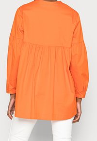 Blouse orange à coupe décontractée, manches longues et corps froncé. Tissu lisse, longueur genou avec un ourlet arrondi.