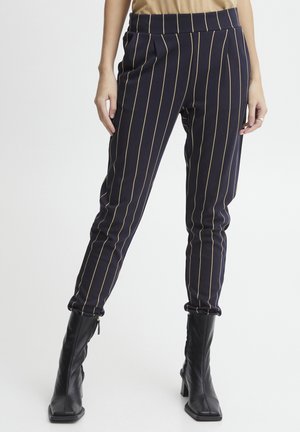 Marineblaue high-waisted Hosen mit vertikalen goldenen Nadelstreifen, elastischem Bund und schmal zulaufenden Beinen, kombiniert mit hohen schwarzen Stiefeln mit Absatz.
