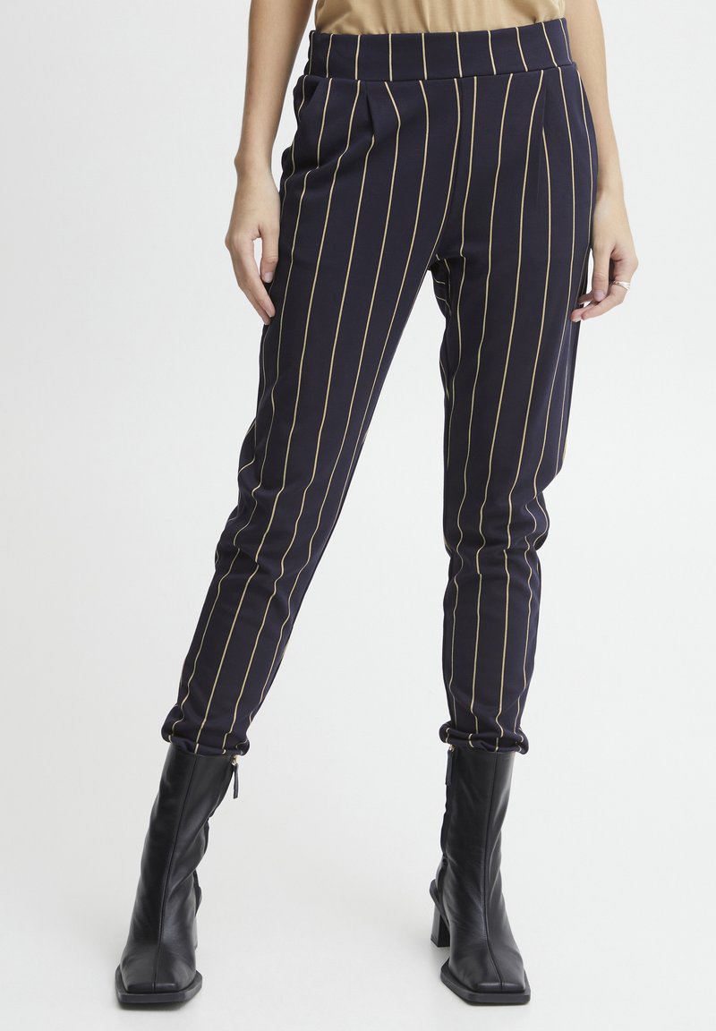 Pantalons marine taille haute avec des rayures dorées verticales, ceinture élastique et jambes fuselées, associés à des bottes noires à talons hauts.