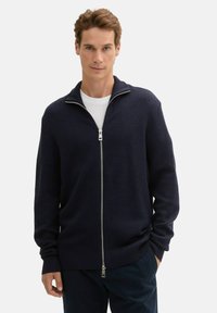 Czarny granatowy kardigan dzianinowy z zamkiem z przodu, wysokim kołnierzem, prążkowanymi mankietami i gładką fakturą, noszony na białej koszulce z okrągłym dekoltem.