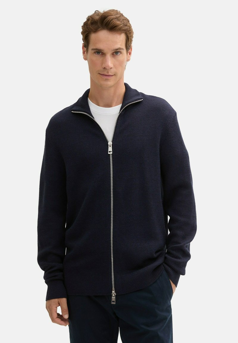 Czarny granatowy kardigan dzianinowy z zamkiem z przodu, wysokim kołnierzem, prążkowanymi mankietami i gładką fakturą, noszony na białej koszulce z okrągłym dekoltem.