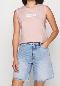 Canotta rosa senza maniche con logo bianco "LEVI'S", abbinata a pantaloni shorts di denim azzurri chiari con orli sfrangiati e una vestibilità dritta classica.