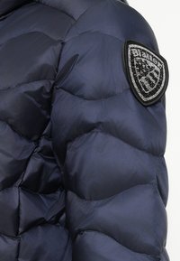 Jūras zila puffa jaka ar quilted dizainu, ar teksturētu audumu un dekoratīvu pleķi ar rhinestones augšējā rokā.