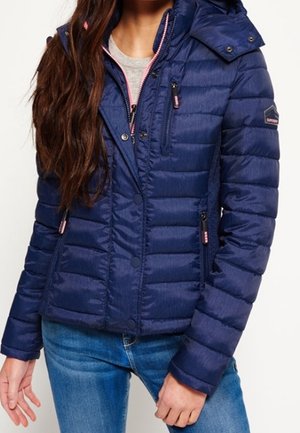 Femme portant une veste matelassée bleu marine avec des poches zippées devant et sur la poitrine, associée à un jean bleu, debout devant un fond uni.