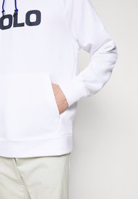 Sudadera blanca hecha de tela suave, con un gran logo negro de "POLO", cordones ajustables azules y un bolsillo tipo canguro.