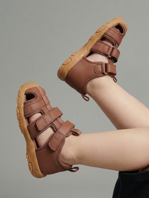 Nogi dziecka w brązowych sandałach zakrywających palce, z podwójnymi paskami na rzep i grubymi gumowymi podeszwami na jednolitym szarym tle.