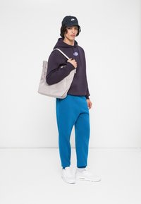 Jovem a usar um boné preto da Nike, um hoodie escuro, calças azul brilhante e ténis brancos, segurando uma grande bolsa de tecido cinza claro no ombro.