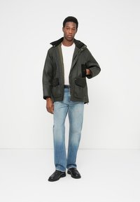 Jachetă parka verde măsliniu cu glugă, având două buzunare la piept, cu fermoar și închidere cu nasturi, purtată peste un tricou alb și blugi bleu deschis.