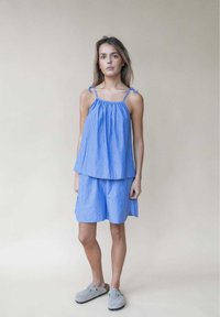 Blau gestreiftes ärmelloses Oberteil mit gerafftem Ausschnitt und verstellbaren Schulterbindungen, kombiniert mit einer passenden Shorts aus demselben Stoff. Graue Clogs.