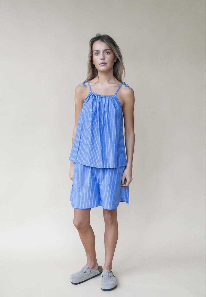 Blau gestreiftes ärmelloses Oberteil mit gerafftem Ausschnitt und verstellbaren Schulterbindungen, kombiniert mit einer passenden Shorts aus demselben Stoff. Graue Clogs.