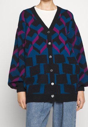 Person trägt eine schwarz geknöpfte Strickjacke mit blauen und violetten geometrischen Mustern über einem weißen Hemd und hellblauen Jeans.