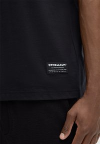 Schwarzes Shirt mit einem Strellson-Etikett am Saum, getragen von einer Person mit dunkler Haut, die Hand entspannt an der Seite, vor einem schlichten Hintergrund.