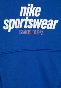 Felpa blu realizzata in tessuto morbido, con grande scritta bianca "nike sportswear" e etichetta arancione "ESTABLISHED 1972". Texture liscia.