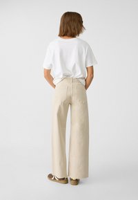 Pantalons larges beiges en tissu lisse, associés à un T-shirt blanc. Poches arrière visibles et taille haute.