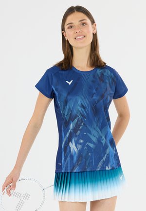 Ung kvinde i blå mønstret sportstrøje og gradient tennisnederdel holder en badmintonketsjer og smiler med en neutral baggrund.