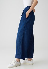 Weite Jeanshose in Dunkelblau, mit elastischem Bund und Kordelzug, seitlichen Taschen und umgeschlagenen Säumen. Kombiniert mit weißen Sneakers.