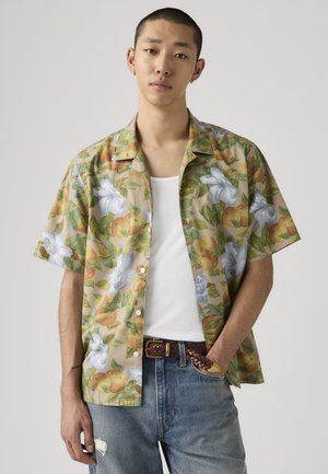 Jeune homme portant une chemise à manches courtes beige à motifs floraux par-dessus un débardeur blanc, avec un jean bleu et une ceinture tressée marron, debout avec une main dans la poche.