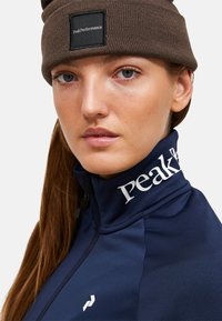 Giacca blu navy con zip e alto colletto, con logo bianco "PeakPerformance" sul colletto. Berretto a maglia marrone con etichetta del logo. Texture liscia.