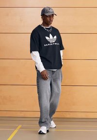 Svart grafisk t-shirt med texten "BASKETBALL", vit långärmad tröja under, grå jeans och vita sneakers. Grå keps kompletterar outfiten.