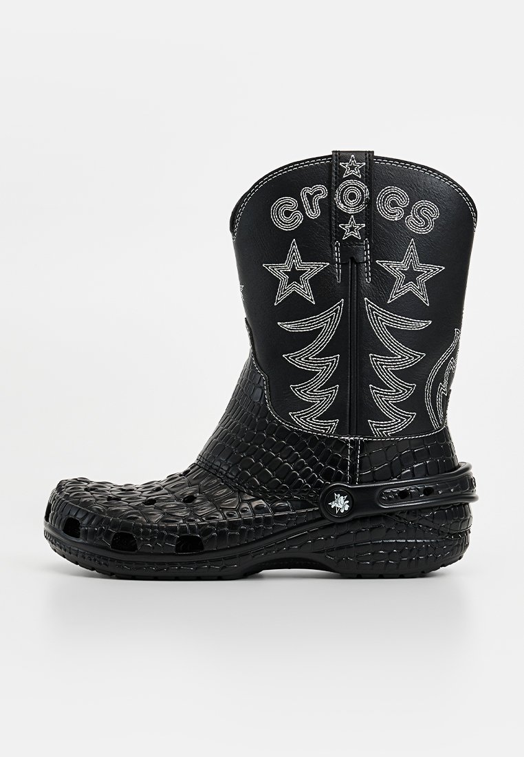 Botas Crocs negras con una superficie texturizada que imita la piel de cocodrilo, diseños en blanco de estrellas y árboles, y una cremallera en la parte posterior para ponérselas y quitárselas fácilmente.