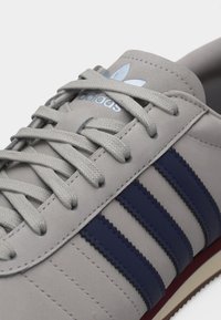 Ανοιχτό γκρι αθλητικό παπούτσι Adidas με μπλε ρίγες, κορδόνια σε ίδιο χρώμα και λογότυπο Adidas στη γλώσσα, εμφανιζόμενο σε κοντινή λεπτομέρεια.