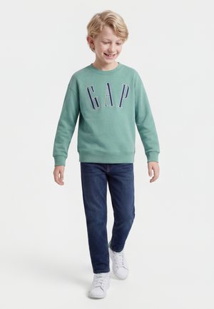 Svetlolasi fant se smehlja in hodi, oblečen v zeleno GAP majico s kapuco, temno modre kavbojke in bele superge na enobarvnem belem ozadju.