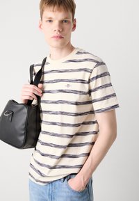 Tricou din bumbac cu dungi bej și linii bleumarin, guler rotund și un mic logo brodat pe piept. Modelul ține o geantă neagră.