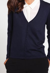 Cardigan bleu marine à manches longues, col en V et petits boutons ; porté sur une chemise blanche à col. Texture douce et coupe ajustée.