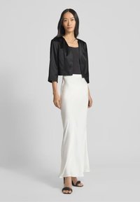 Giacca cropped in satin nero su un top nero, abbinata a una lunga gonna in satin bianco. La modella indossa sandali con tacco neri e si trova su uno sfondo neutro.
