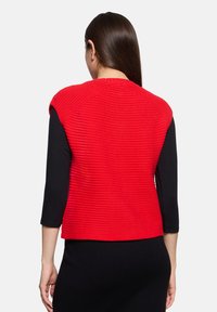 Czerwona dzianinowa kamizelka z teksturowanym wzorem, bez rękawów, z okrągłym dekoltem, noszona na czarnej bluzce z długim rękawem.