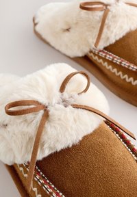 Chaussons en daim marron avec doublure blanche duveteuse et coutures décoratives, noués avec des lacets en daim marron en nœud.
