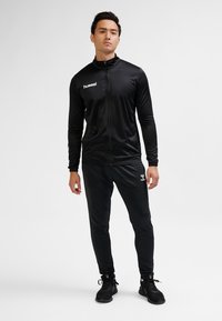 Black athletic tracksuit featuring a zip-up jacket and slim-fit pants, made of smooth fabric with a logo on the chest and leg.
