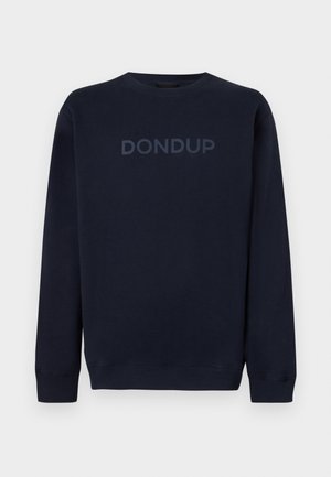 Homme à la tête rasée, debout, les mains dans les poches, portant un sweat-shirt noir à col rond avec le logo « DONDUP », jean foncé et baskets marron sur fond blanc.