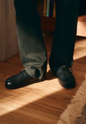 Schwarze Gummi-Clogs mit Schnalle, die mit strukturierten Mustern auf der Oberseite versehen sind. Getragen mit grünen Hosen, auf einem Holzboden.