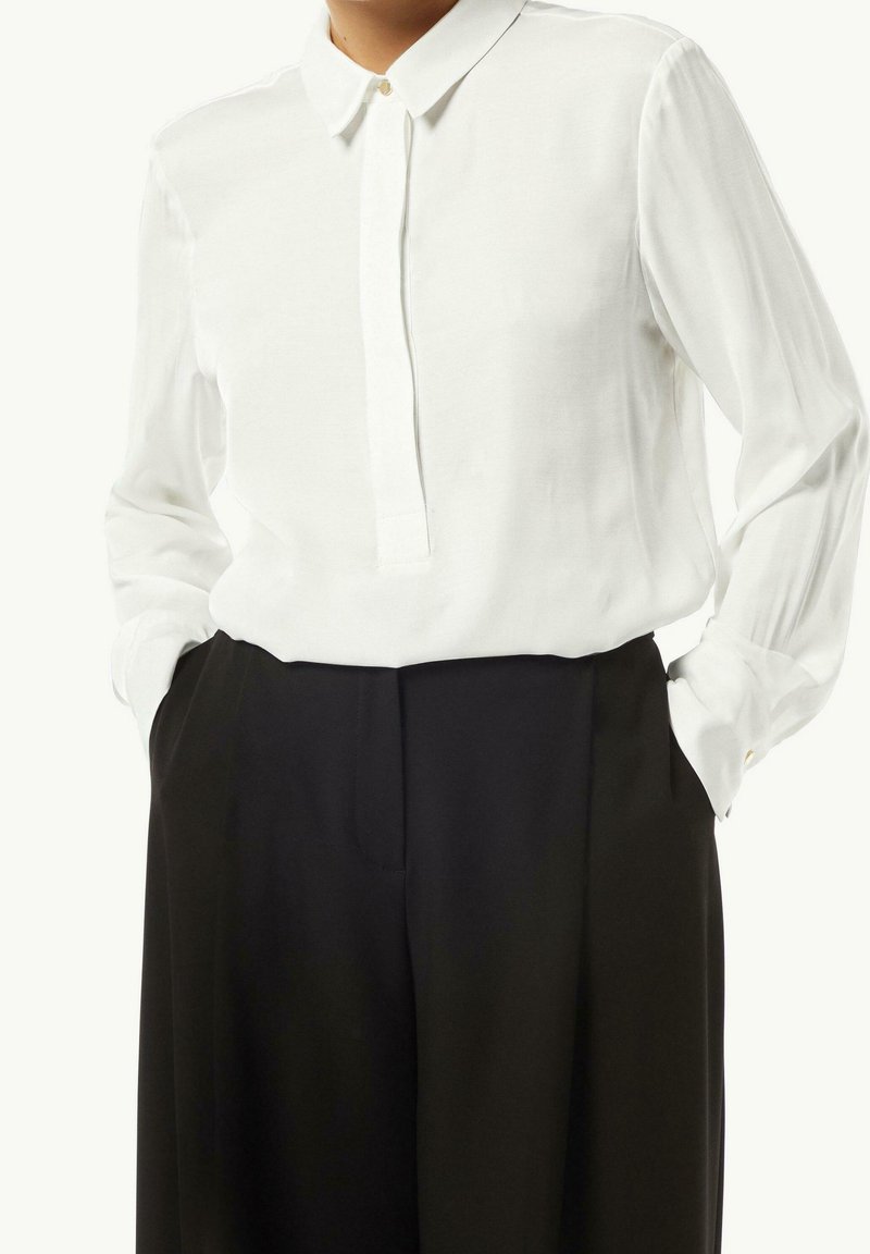 Camicia bianca a maniche lunghe con colletto, caratterizzata da una sottile pattina dei bottoni, abbinata a pantaloni neri a vita alta con tasche laterali.