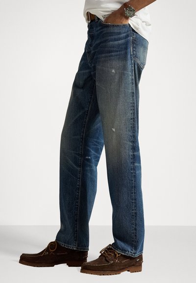 Man wearing faded blue denim jeans, brown leather belt, white t-shirt, silver wristwatch, and brown suede lace-up shoes standing sideways.