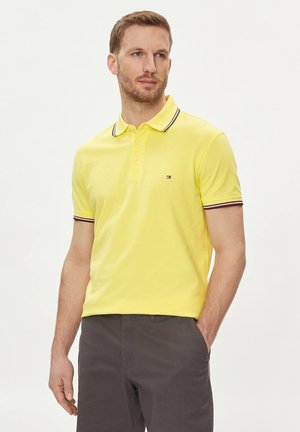Polo giallo con rifiniture nere e rosse su colletto e maniche, con un piccolo logo sul petto. Indossato con pantaloni corti grigio scuro.