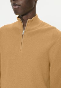 Homme portant un pull demi-fermeture éclair texturé couleur chameau, fermeture éclair partiellement fermée, sur un fond blanc uni.