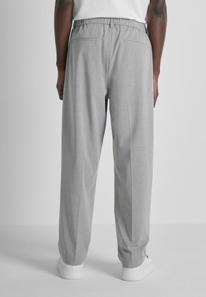 Uomo con pantaloni plissettati grigio chiaro e sneakers bianche, in piedi contro uno sfondo chiaro e uniforme, rivolto di spalle alla fotocamera.