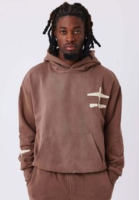 Felpa con cappuccio marrone, dotata di tasca frontale, con un design astratto di un beige chiaro sul petto e sul maniche sinistra; tessuto morbido e testurizzato.