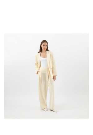 Kobieta ubrana w kremowy garnitur z szerokimi nogawkami i marynarką, pod którą ma biały top, stojąca na jednolitym białym tle.