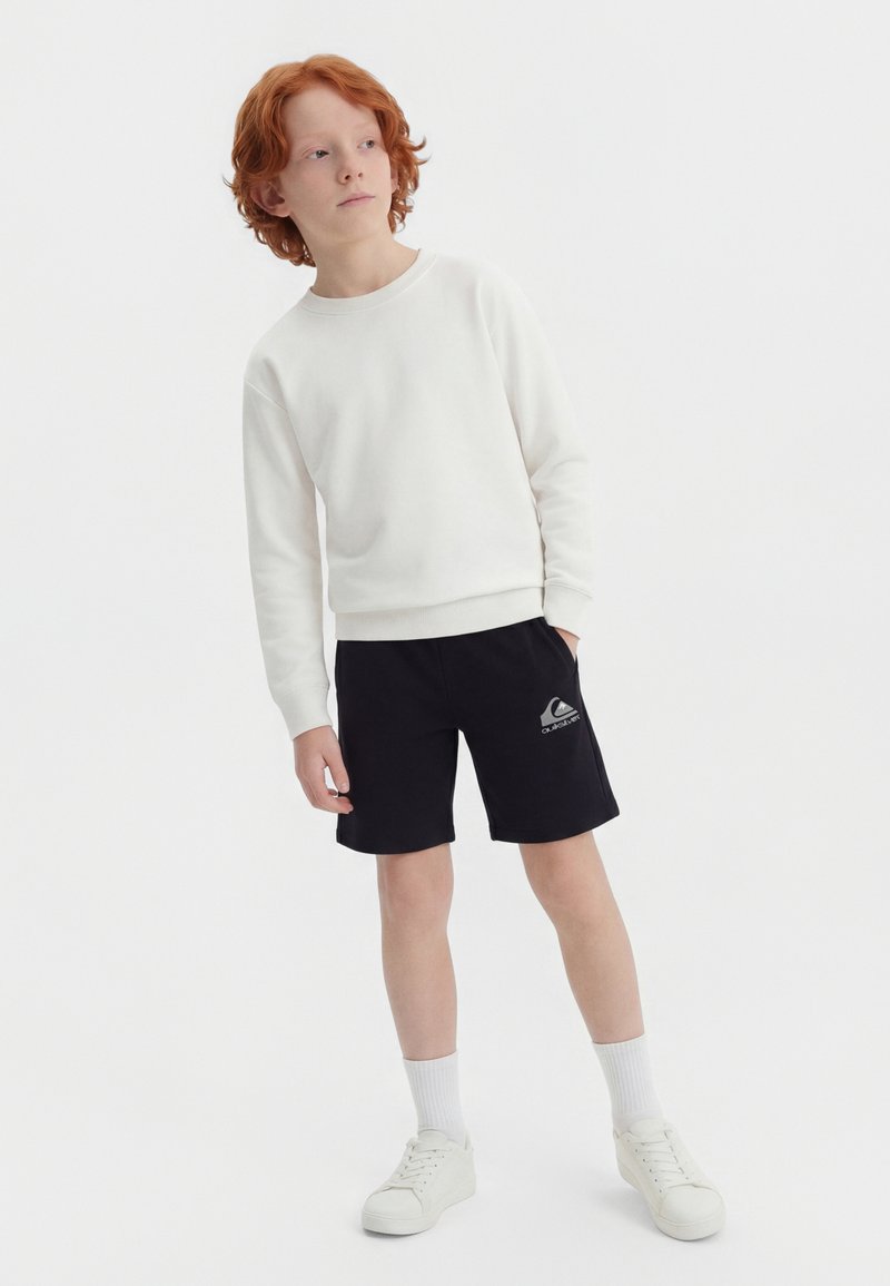 Chłopiec o rudych włosach, ubrany w białą bluzę, czarne spodenki Quiksilver, białe skarpetki i białe buty sportowe, stoi z jedną ręką w kieszeni.