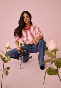 Pikkade tumedate juustega naine, kes kannab punasevalgete triipudega särki ja pükse, küürutab, hoides roosat roosi klaasvases roosal taustal.