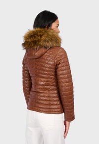 Bruine, gewatteerde leren jas met een grote faux-fur capuchon. Heeft een getailleerde pasvorm en een soepele textuur, gecombineerd met witte broeken.